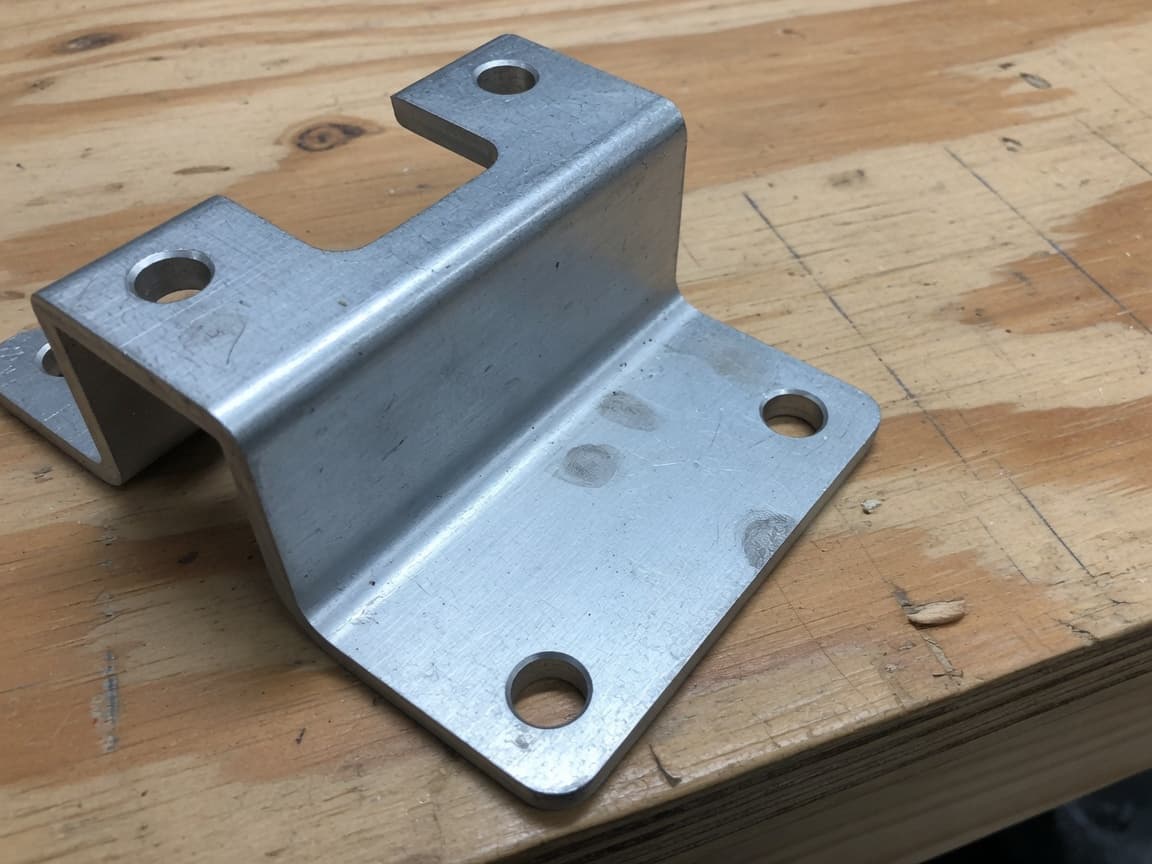 Formed aluminum bracket with mounting holes on a plywood bench.